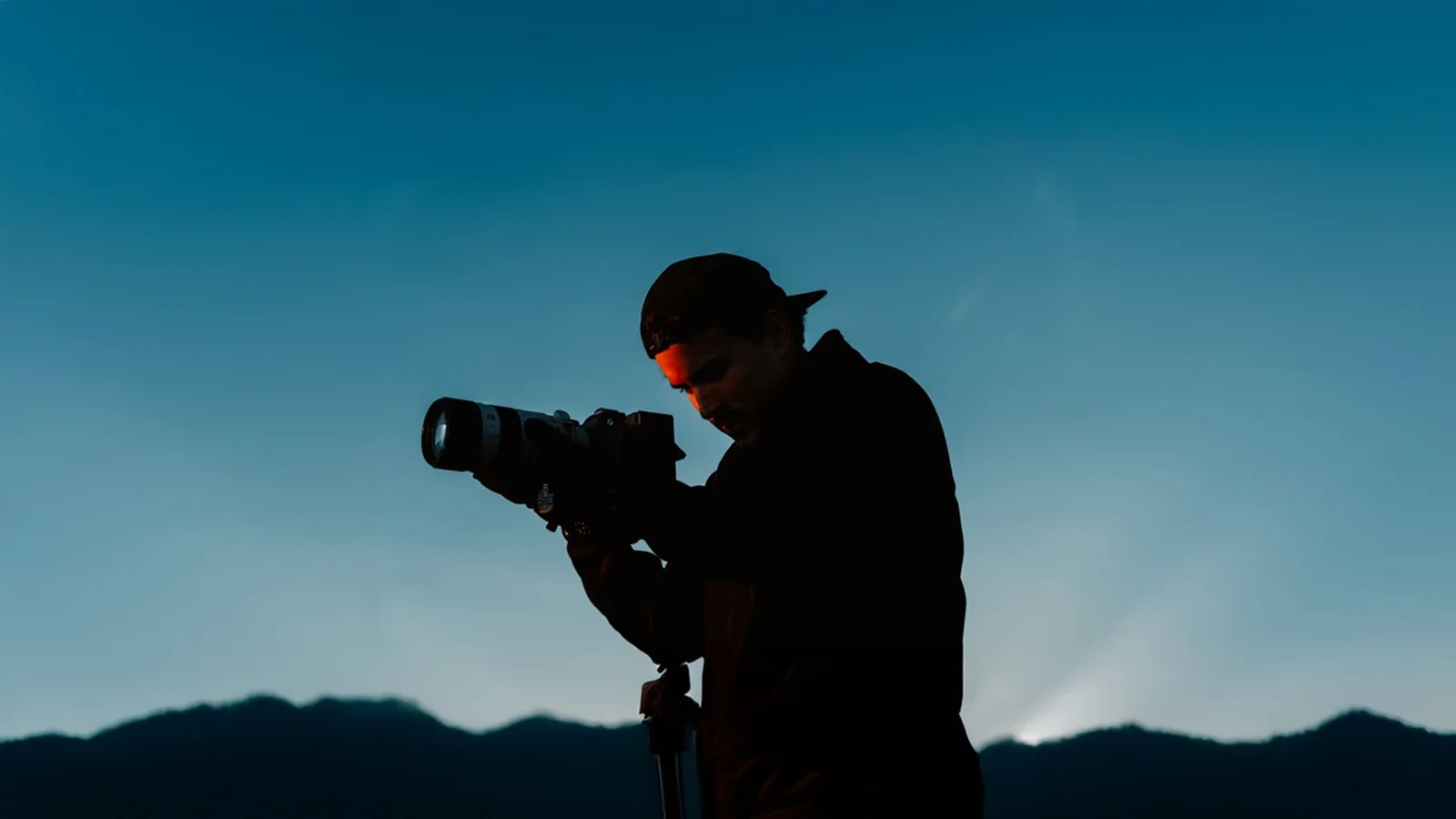 Vidéaste en silhouette à contre-jour, caméra sur trépied, face aux crêtes