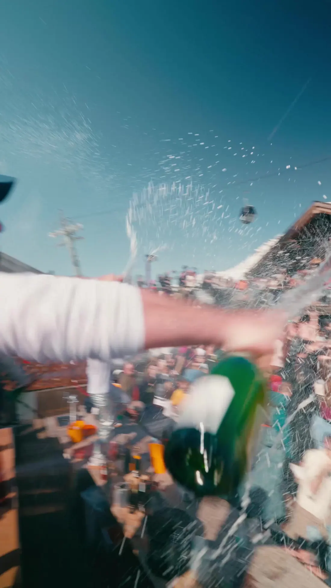 Bouteille de champagne qui jaillit vers la caméra au-dessus de la foule, ciel bleu d'altitude