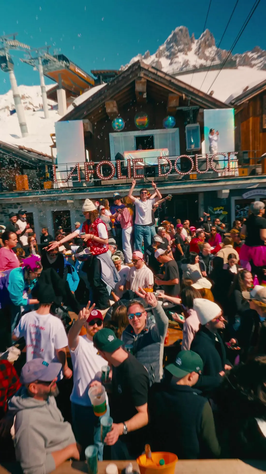 Façade en bois de la Folie Douce avec son enseigne, foule les bras levés et sommet rocheux derrière, closing d'avril 2026