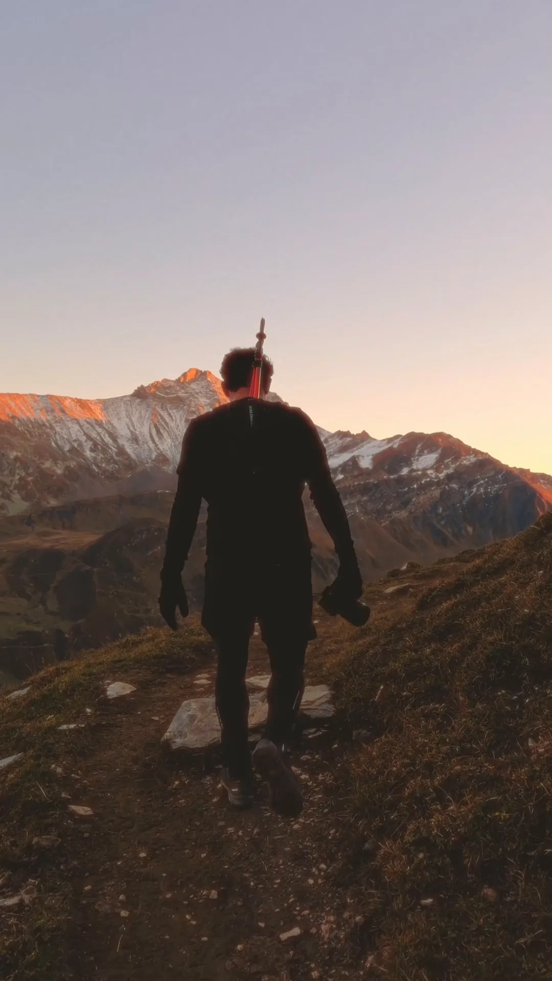 Vidéaste de dos sur un sentier de crête, trépied sur l'épaule, sommets enneigés éclairés par une lumière rasante