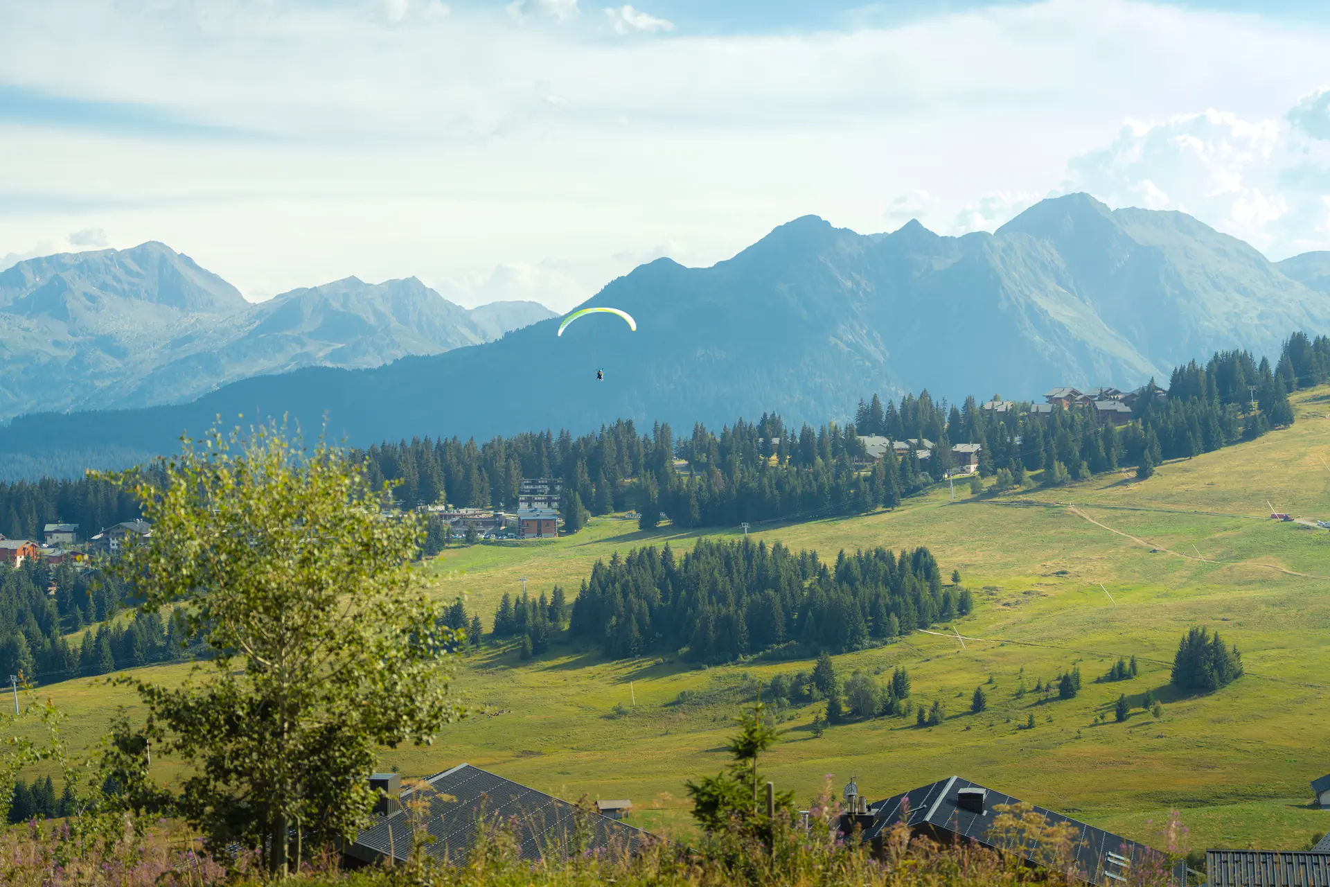 Alpages et chalets des Saisies au premier plan, parapente dans le ciel et massif du Mont-Blanc à l'horizon