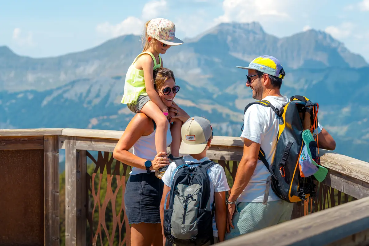 Famille accoudée à la rambarde d'un belvédère en bois, enfant sur les épaules, montagnes en face