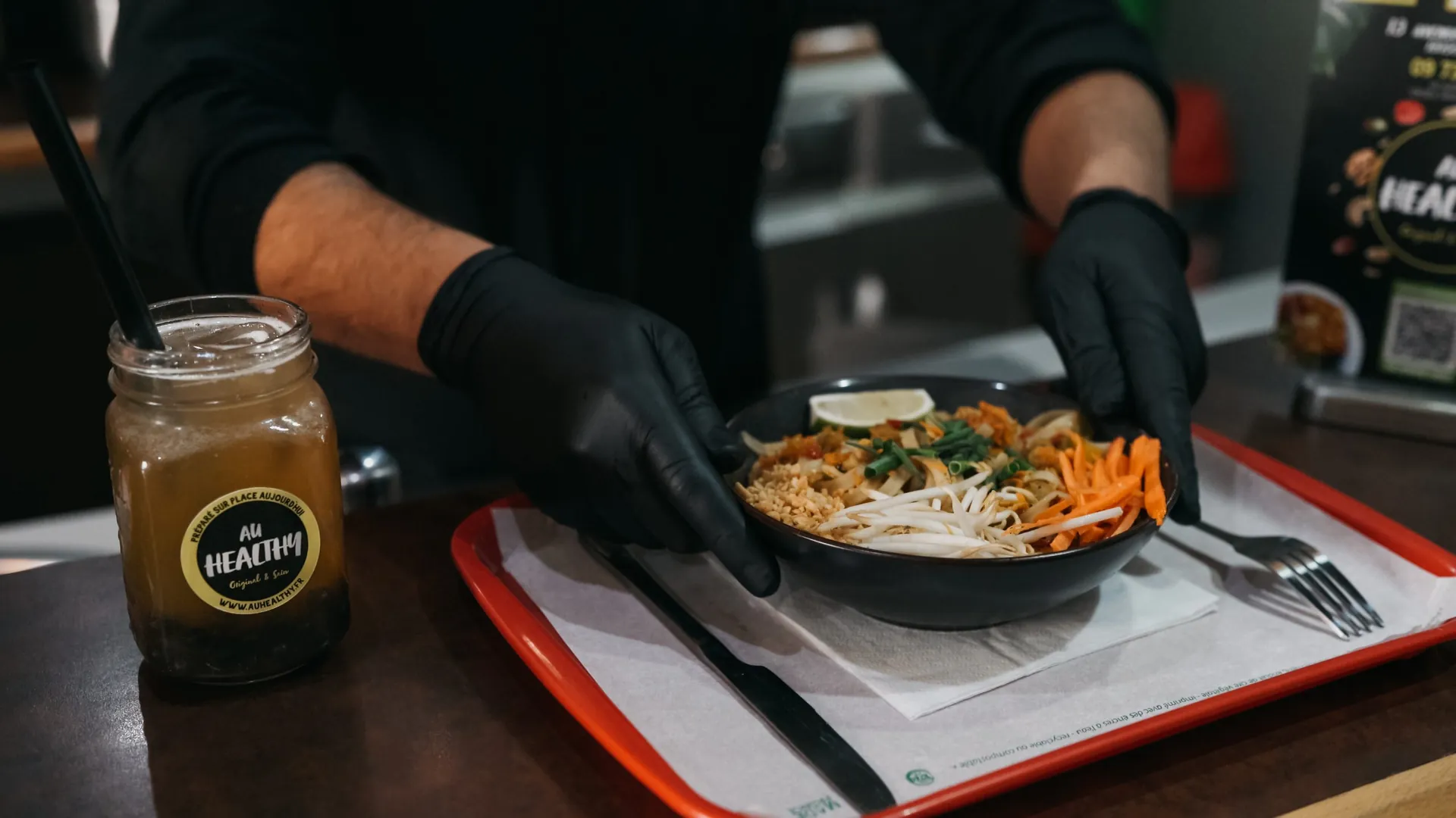 Plan du film Au Healthy : des mains gantées posent un bowl de nouilles sur le plateau, pot de sauce maison au premier plan