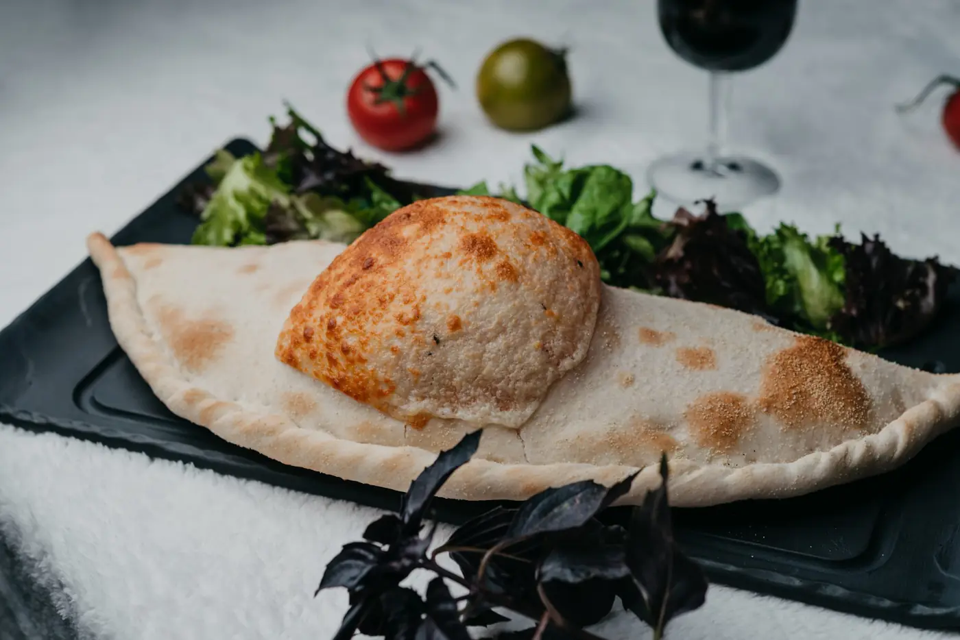 Calzone dorée servie sur ardoise avec salade verte, tomates et verre de vin derrière