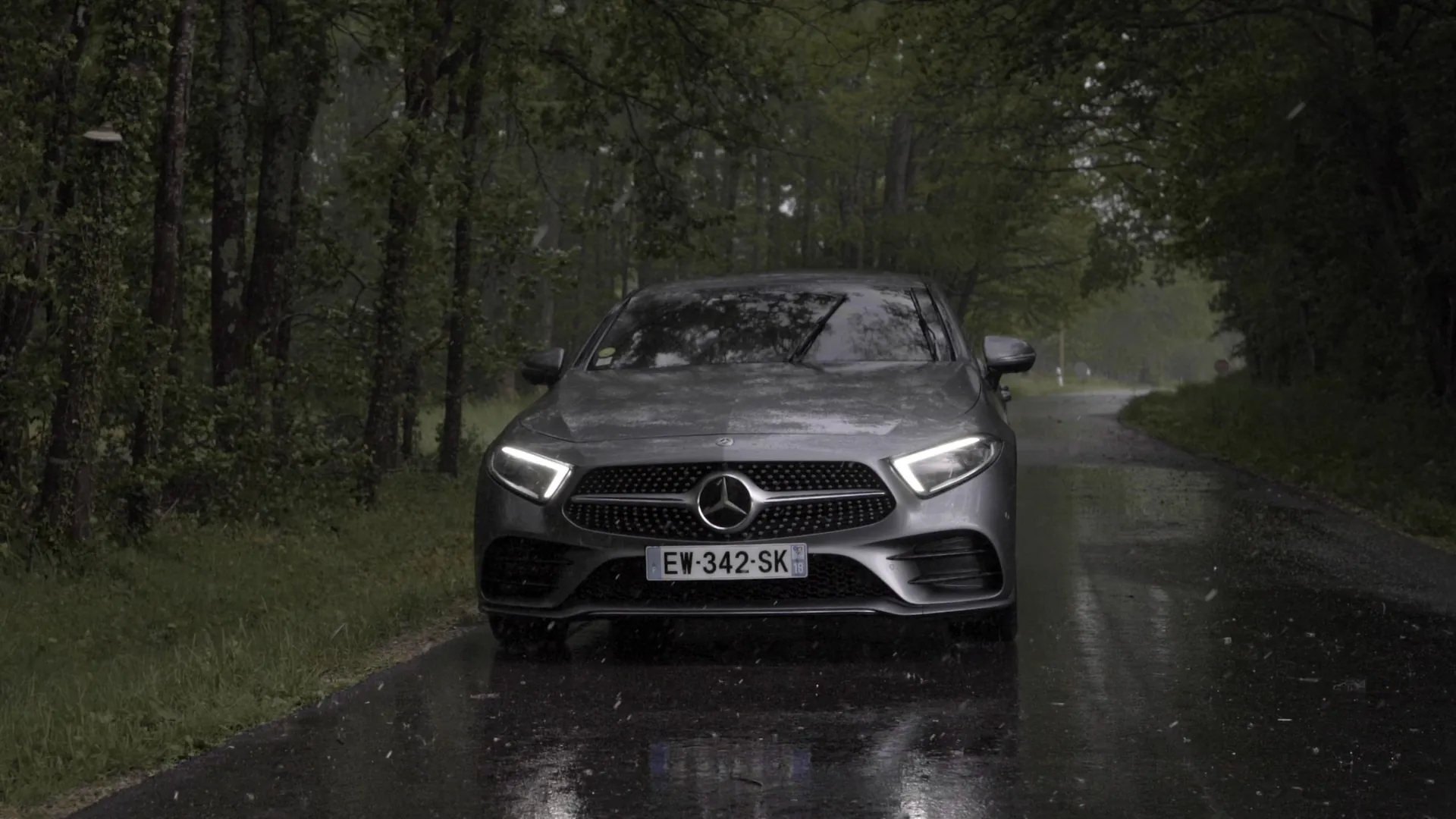 Mercedes CLS AMG Line grise phares allumés sur une route forestière détrempée, sous la pluie