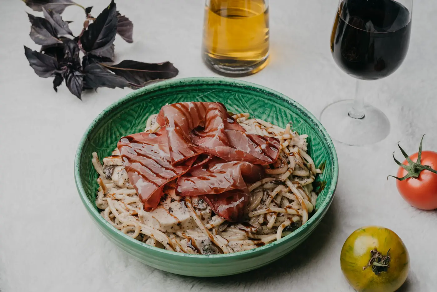 Assiette de pâtes au jambon cru dans un bol vert, verres de vin et tomates autour