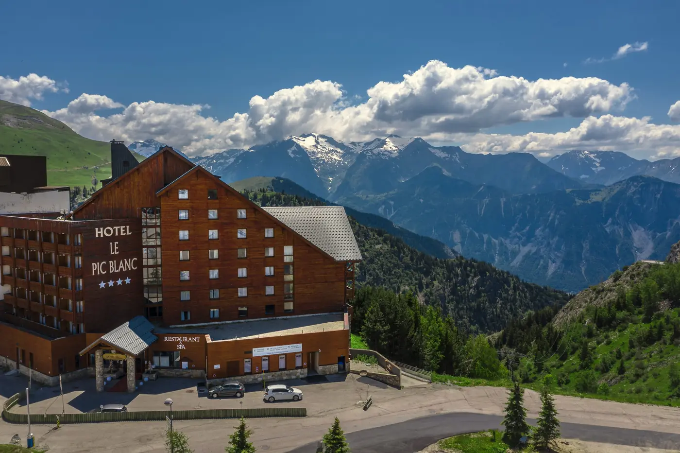 Vue aérienne de l'hôtel Le Pic Blanc à l'Alpe d'Huez, façade en bois et montagnes enneigées derrière