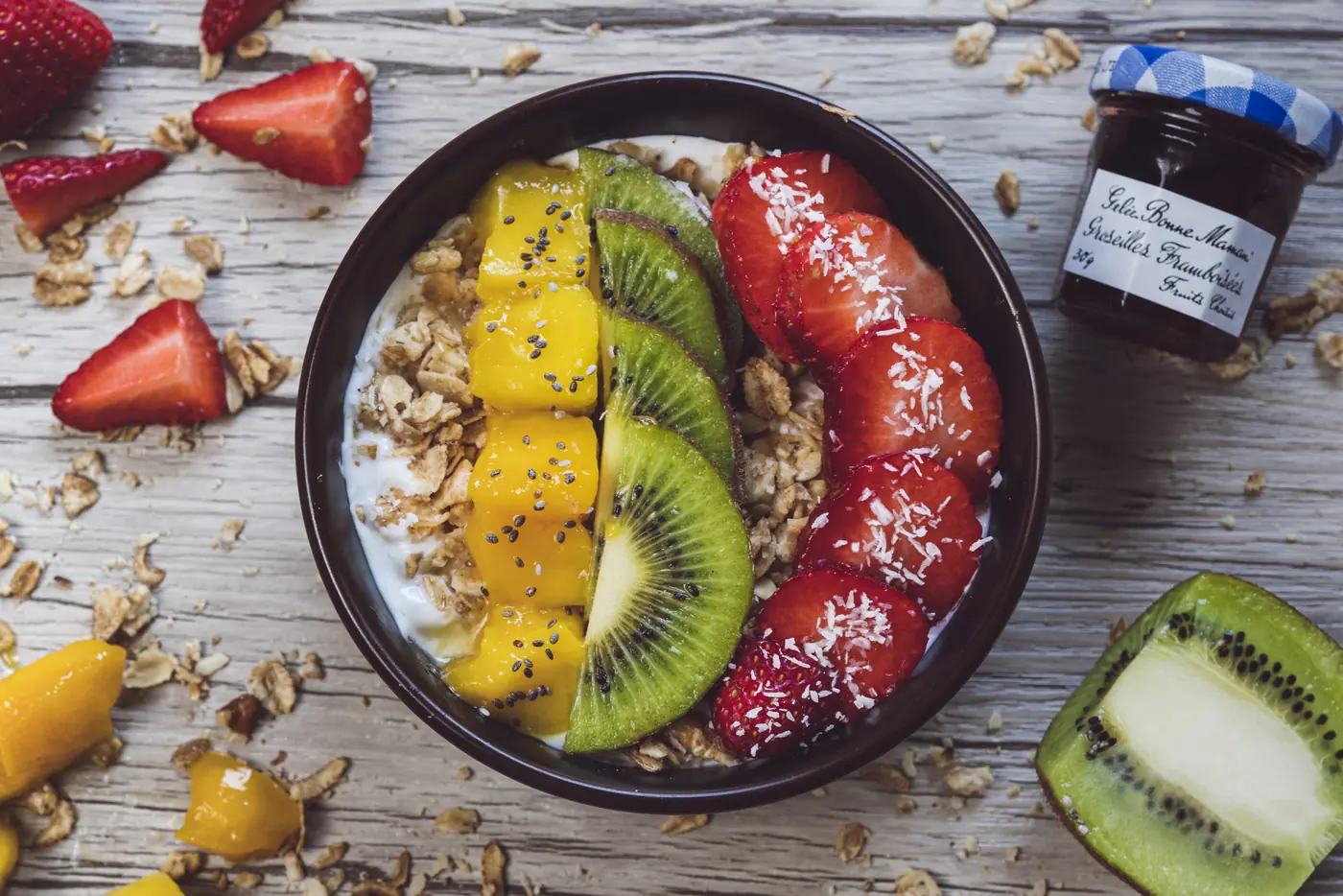 Bowl de granola, mangue, kiwi et fraises vu du dessus, petit pot de confiture à côté