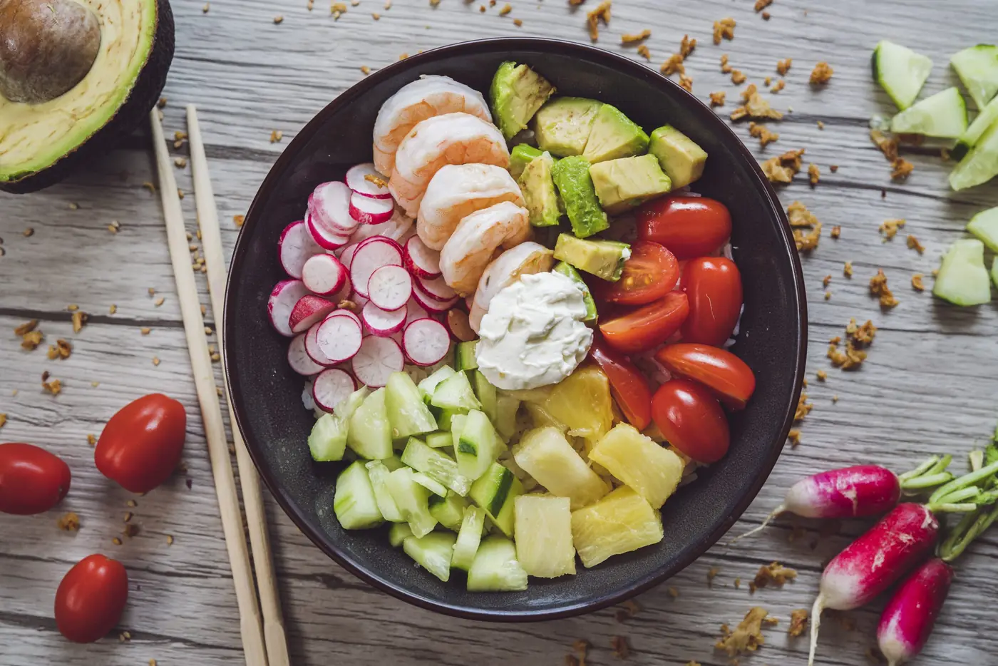 Poke bowl aux crevettes, radis, concombre, avocat et ananas, photographié du dessus sur une table en bois