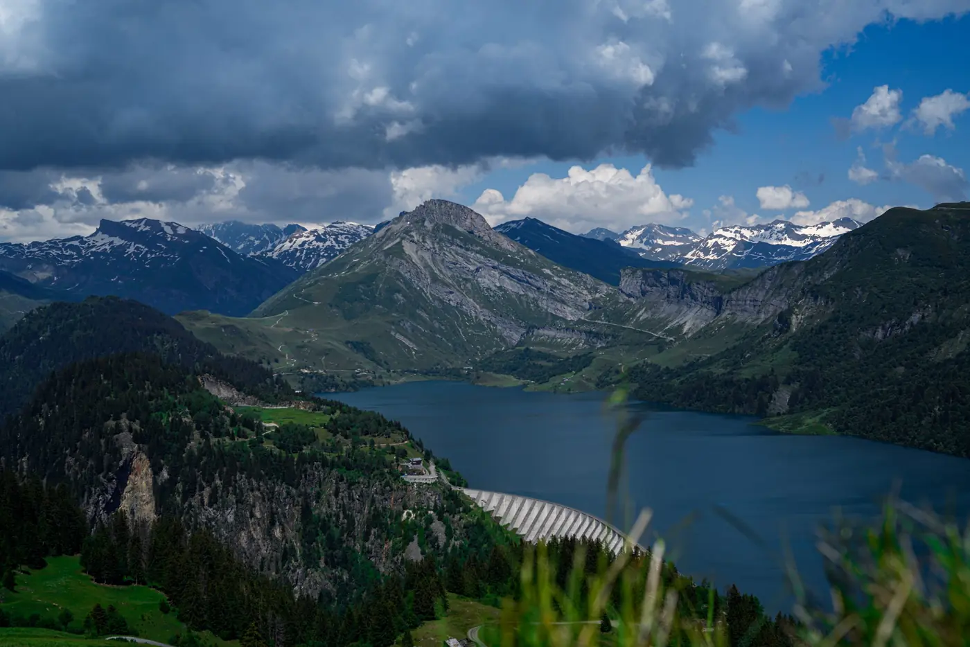 Lac de barrage turquoise entre des versants boisés, sommets encore enneigés en juin