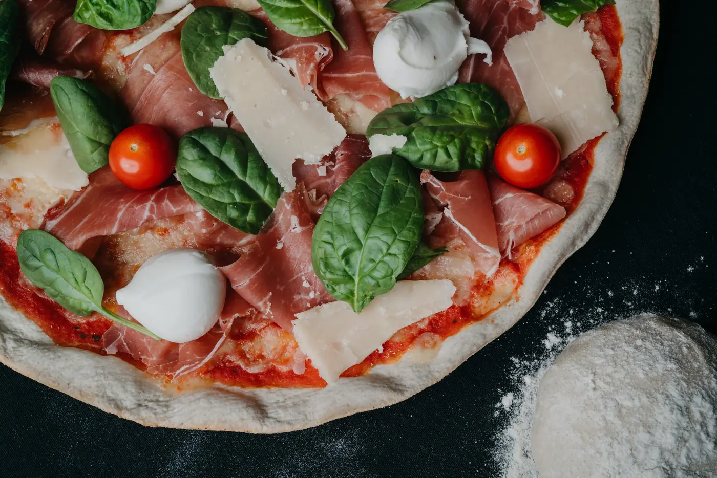 Pizza au jambon cru, tomates cerises, mozzarella et copeaux de parmesan, photographiée de près sur fond sombre