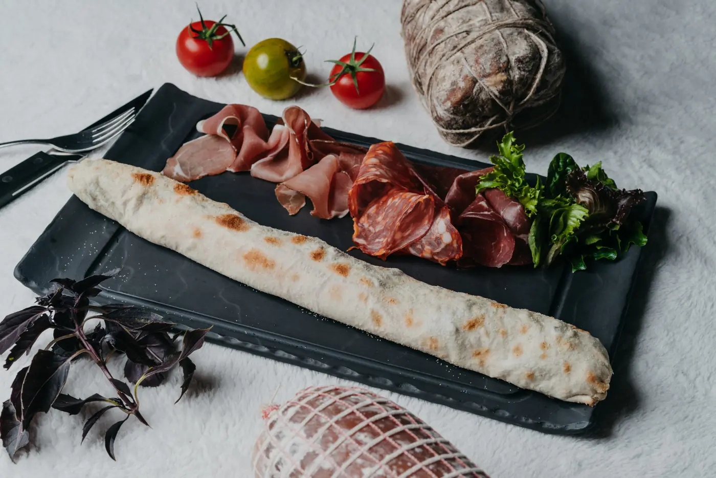 Pizza longue garnie de charcuterie et de salade sur une ardoise, salami et tomates autour