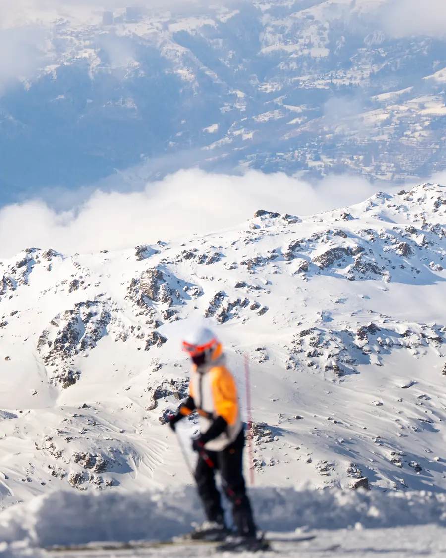 Skieur seul au pied d'un versant rocheux enneigé, vallée et nuages en contrebas, Val Thorens