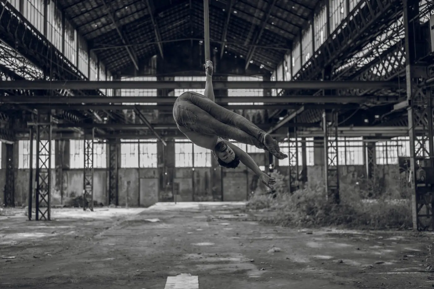 Artiste aérien replié sur lui-même dans la halle désaffectée, végétation qui reprend le sol