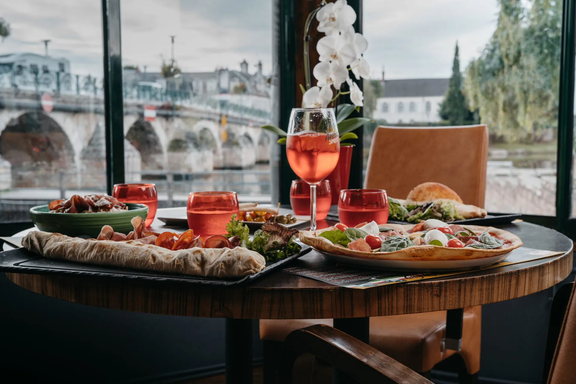 Table dressée avec pizzas, salades et cocktails devant la baie vitrée, pont de pierre en arrière-plan